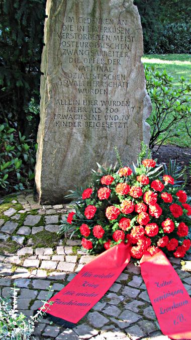 Gedenkstein und Blumengebinde mit Schleifen: Nie wieder Krieg. Nie wieder Faschismus! Kulturvereinigung Leverkusen e.V.