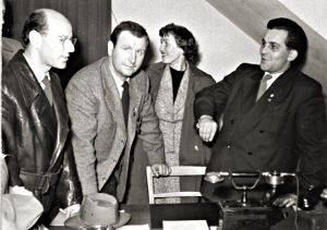 Vier-Personen-Gruppe vor Schreibtisch in engem Zimmer.