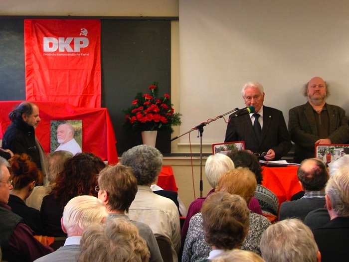 Günter Judick am Rednerpult im Saal der Kulturvereinigung Leverkusen. Im Hintergrund ein Bild von Manni Demmer.